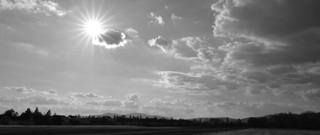 Panoramaaufnahme in schwarz-weiß, am unteren Arnd sind die Silhouetten von Bäumen und Häusern in der Ferne sichtbar, darüber ein weitläufiger Himmel mit Haufenwolken in der Ferne und im rechten Bildbereich, im oberen linken Drittel die Sonnen mit einem 18-zackigen Sonnenstern