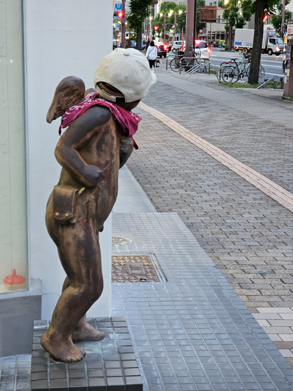 Bronzefigur eines Engels mit einer Basball-Kappe und einem Halstuch bekleidet schaut neugierig um eine Ecke