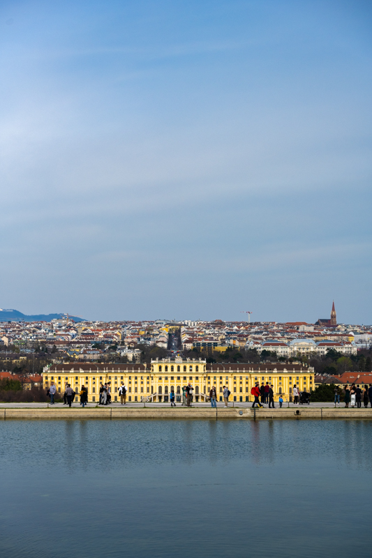 Ein langgezogenes, älteres gelbes Gebäude, im Vordergrund spazierende Menschen und ein Gewässer, der Himmel darüber macht den Großteil des Bildes aus