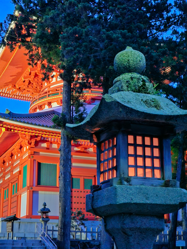 Teil eines roten, japanischen Tempels, davor eine Laterne mit rotem Licht, dazwischen ein Baum, die Dämmerung hat gerade begonnen