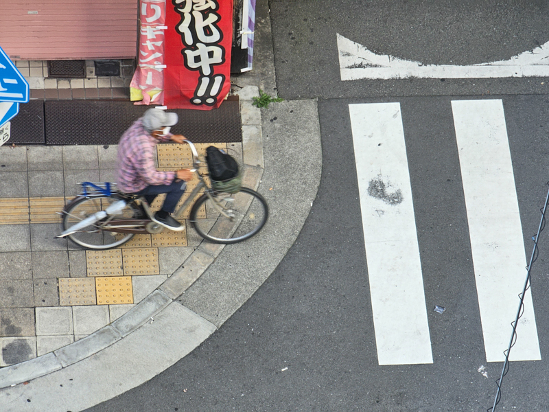 Aufnahme aus der Vogelperspektive, ein Radfahrer überquert eine Straße, japanische Schriftzeichen sind an einer Hauswand sichtbar