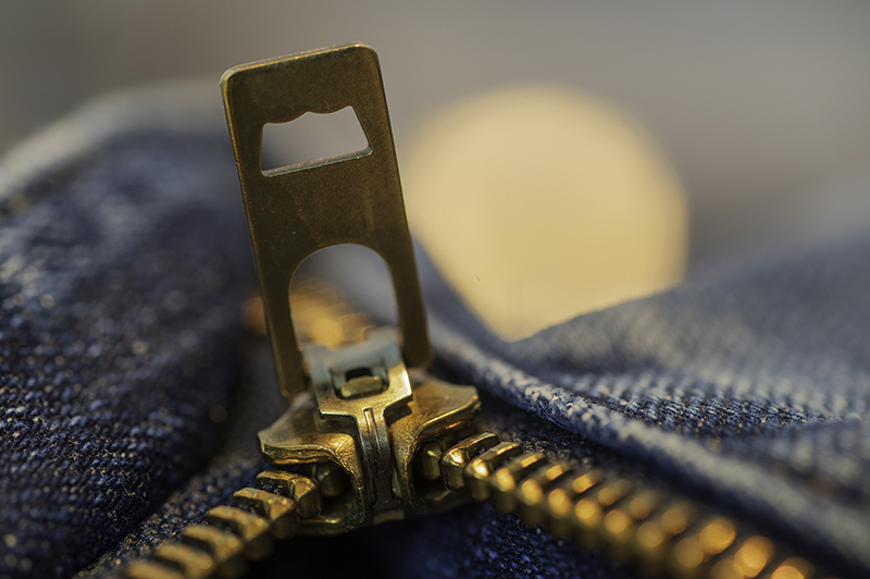Nahaufnahme eines bronzefarbenen Zippverschluss-Schlittens eines Kleidungstücks aus Jeansstoff, dahinter ein unscharfer goldfarbener Kreis