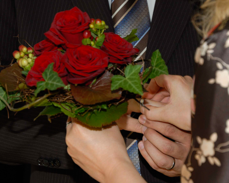 Anstecken eines Rings bei einer Hochzeit mit einer Hand, in der anderen Hand wird ein kleiner Blumenstrauß mit roten Rosen gehalten