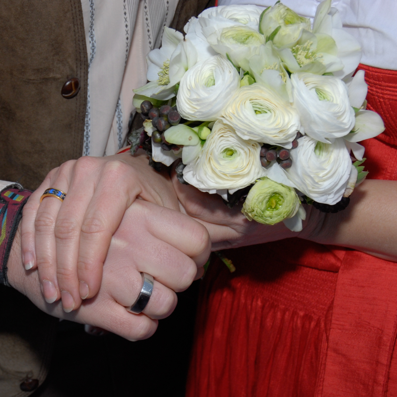 Eine Frauenhand umfasst eine Männerhand, beide haben ein Ring am Ringfinger - in der anderen Han der Frau ein Brautstrauß mit weißen Rosen
