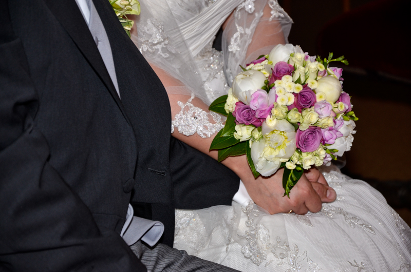 Elegant gekleideter Mann hält die Hand einer Frau im Brautkleid - beide sitzen - in der anderen Hand hält die Frau einen bunten Brautstrauß